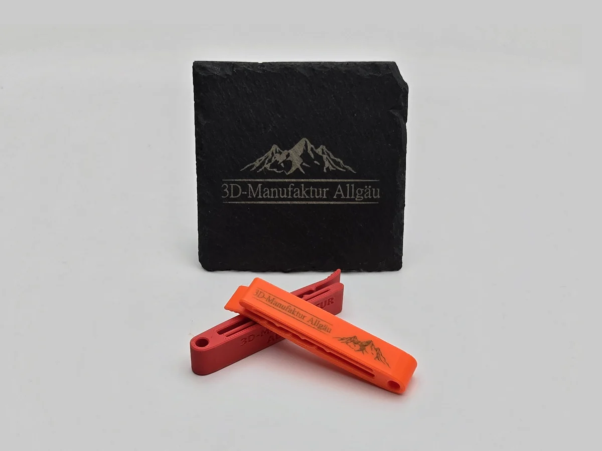 Kroko Kartenhalter in Neon Orange mit gravierter Aufschrift 3D-Manufaktur Allgäu und Bergmotiv, dahinter einer in Rot mit eingeprägtem Schriftzug